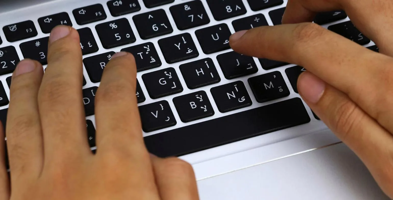 Hands typing on a laptop keyboard with bilingual Arabic and English key labels