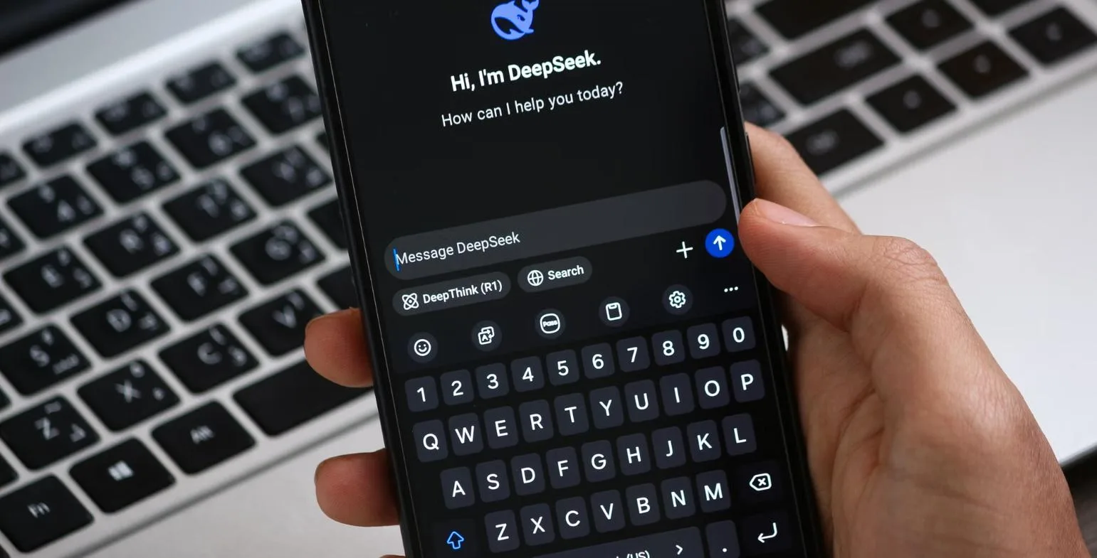 Hand holding a smartphone with the DeepSeek AI assistant open above a laptop keyboard