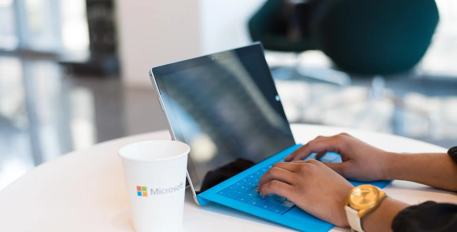 Hands typing on a Microsoft Surface tablet with a blue keyboard beside a Microsoft cup