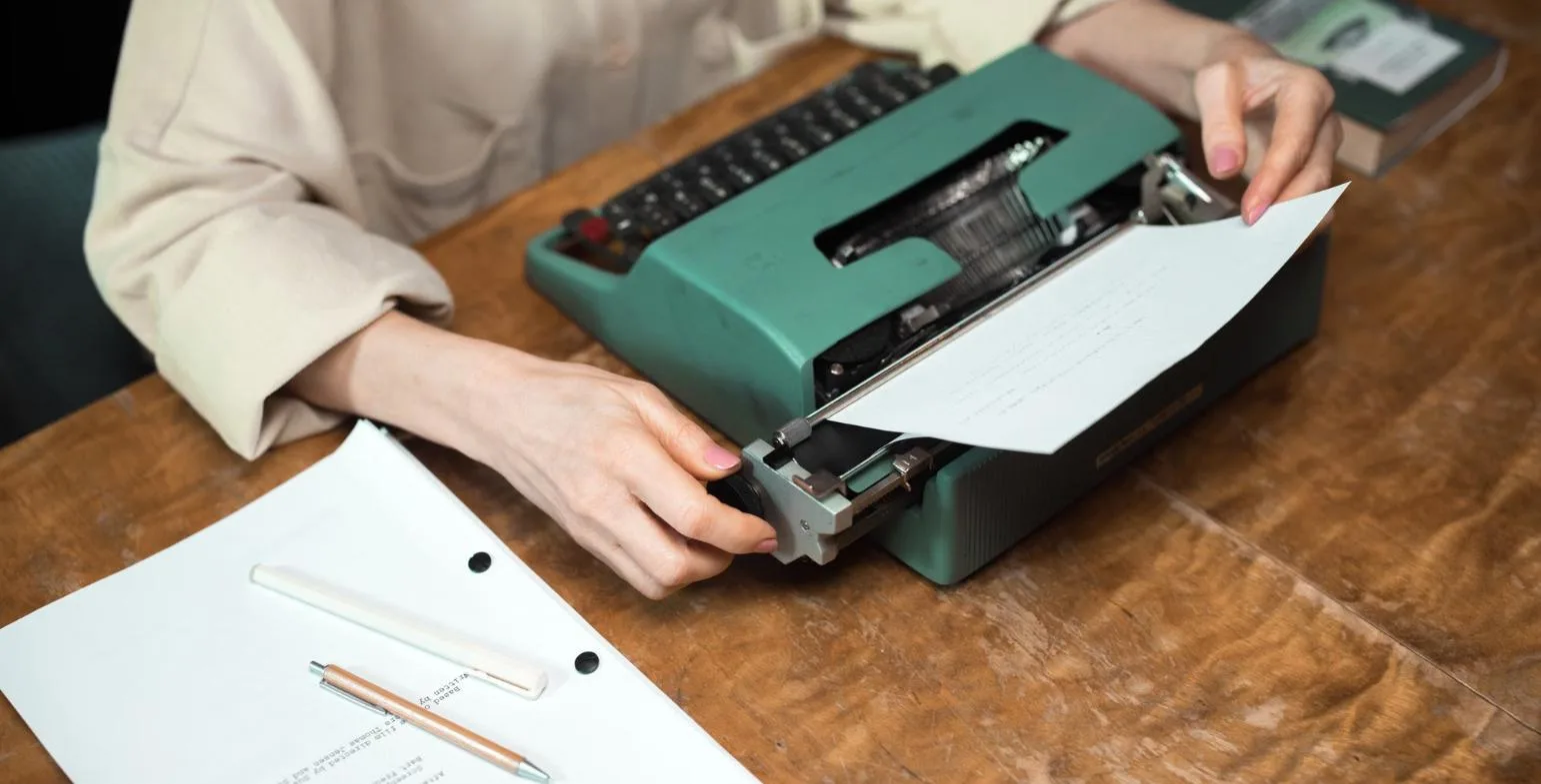 Writer feeding a sheet of paper into a typewriter beside printed pages