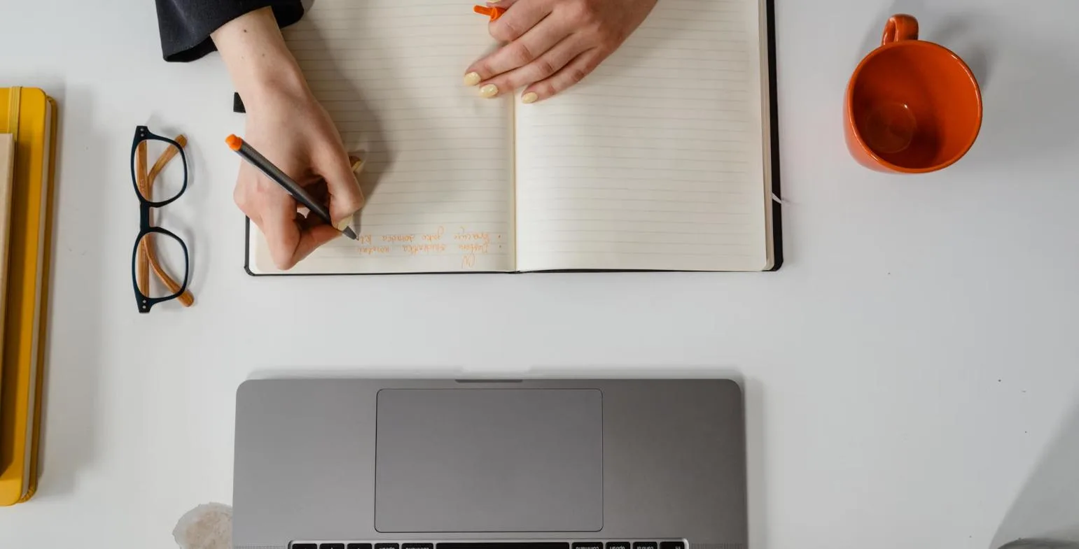 Overhead view of a person writing in an open notebook beside a laptop and coffee cup