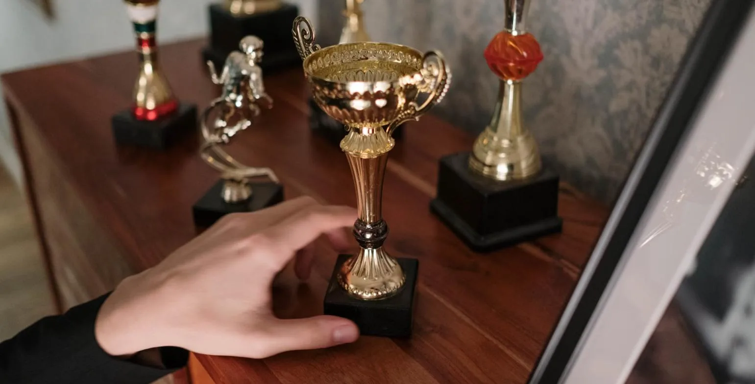 Row of gold trophies displayed on a wooden shelf as a hand places one down
