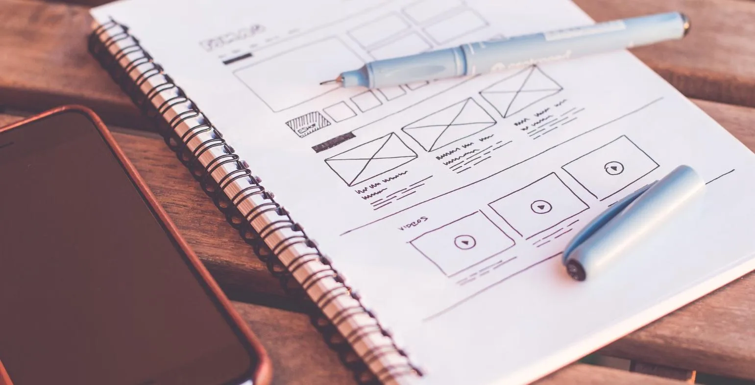 Hand-drawn website wireframe sketch in a notebook beside a phone and pen