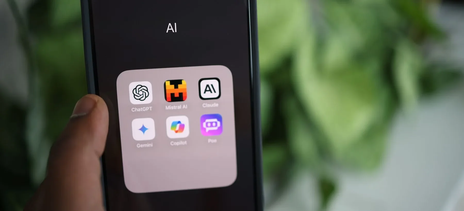 Hand holding a phone showing a folder of AI assistants including ChatGPT, Gemini, Claude, Copilot, Mistral and Poe